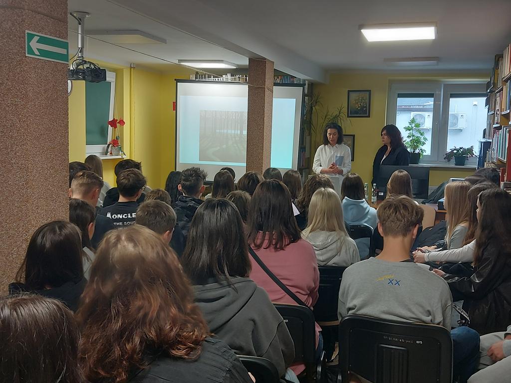 Dwie kobiety stoją przed siedząca grupą młodzieży. Obok kobiet jest ekran rzutnika. Za kobietami jest odsłonięte okno. Po prawej stronie są regały z książkami.
