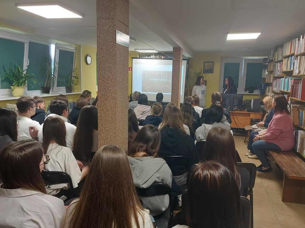 Dwie kobiety stoją przed siedząca grupą młodzieży. Obok kobiet jest ekran rzutnika. Za kobietami jest zasłonięte okno. Po prawej stronie są regały z książkami.