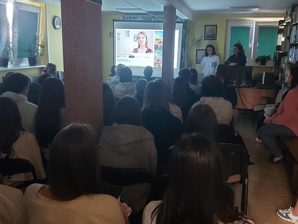 Dwie kobiety stoją przed siedząca grupą młodzieży. Obok kobiet jest ekran rzutnika, na którym jest puszczony film. Za kobietami jest okno. Po prawej stronie są regały z książkami. Po lewej stronie jest zasłonięte okno.