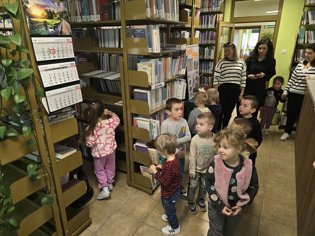 Na środku wypożyczalni stoi grupa dzieci, za nimi trzy panie nauczycielki. Po lewej stronie stoją regały z książkami.