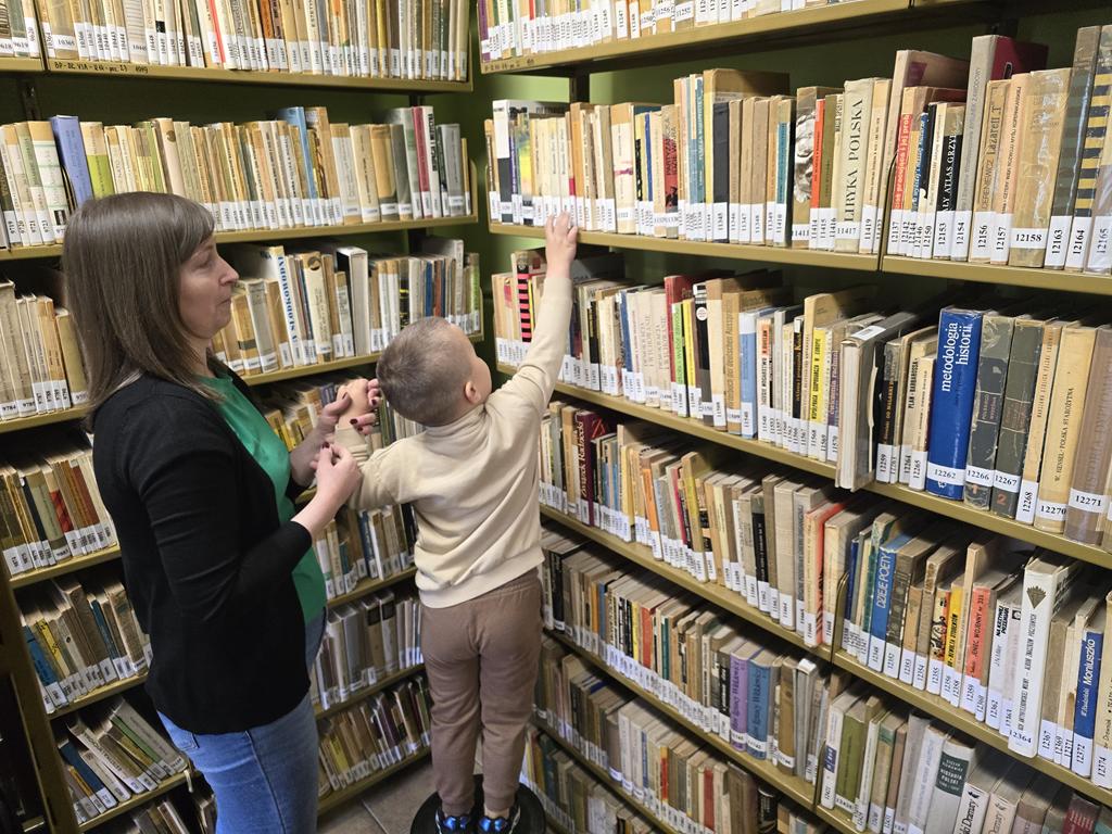 Bibliotekarka w zielonej bluzce, czarnym swetrze i niebieskich spodniach trzyma dziecko w brązowych spodniach i beżowej bluzce za rękę, gdy wchodzi na stołek, aby dosięgnąć do jak najwyższej półki. W tle regały z książkami.