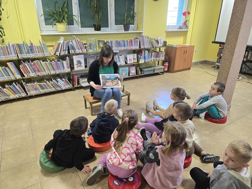 Bibliotekarka w zielonej bluzce, czarnym swetrze i niebieskich spodniach siedzi na stołku i pokazuje obrazek z etapami powstania papieru. Przed nią siedzą na siedziskach przedszkolaki w kolorowych ubraniach. Za bibliotekarką stoją regały z książkami.