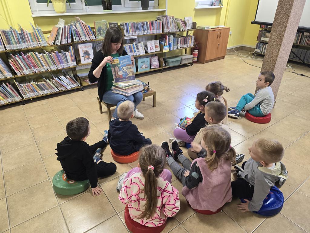 Bibliotekarka w zielonej bluzce, czarnym swetrze i niebieskich spodniach siedzi na stołku i pokazuje różne formy wydania poszczególnych książek. Przed nią siedzą na siedziskach przedszkolaki w kolorowych ubraniach. Za bibliotekarką stoją regały z książkami.