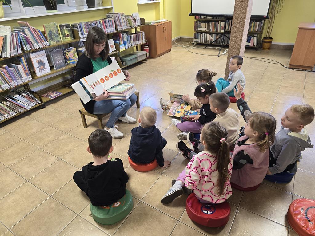Bibliotekarka w zielonej bluzce, czarnym swetrze i niebieskich spodniach siedzi na stołku i pokazuje różne formy wydania poszczególnych książek. Przed nią siedzą na siedziskach przedszkolaki w kolorowych ubraniach i oglądają książki. Za bibliotekarką stoją regały z książkami.