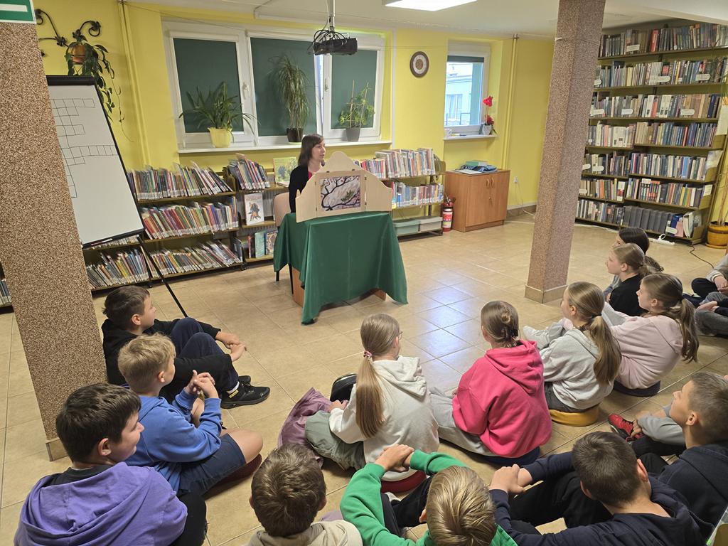 Nauczyciel bibliotekarz w czarnym swetrze i różowej bluzce siedzi przy stoliku. Na stoliku stoi teatrzyk kamishibai, w którym jest umieszczone opowiadanie "Trzy siostry". Po prawej stronie od bibliotekarza stoi tablica do pisania z krzyżówką. Przed Bibliotekarzem siedzi grupa dzieci w kolorowych ubraniach. W tle widać regały z książkami.
