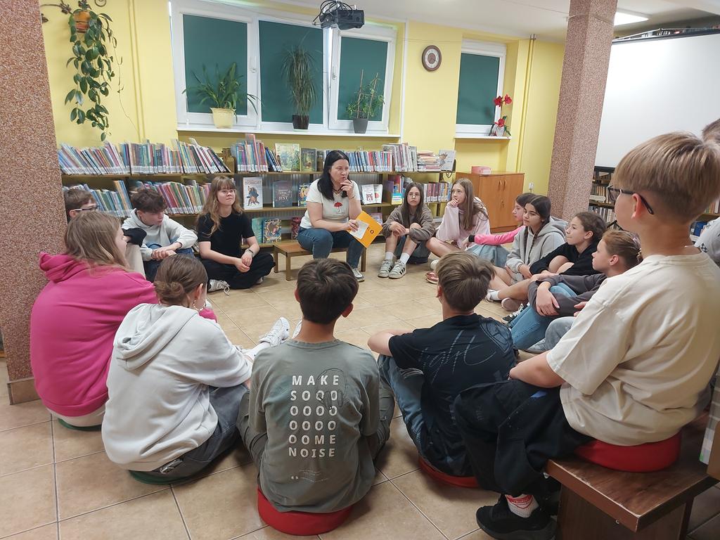 Grupa dzieci 12/13-letnich siedzi w kręgu na podłodze i patrzy na prowadzącą zajęcia. Ta również siedzi w kręgu. Jest ubrana w białą podkoszulkę z motywem Muminków, głowę wspiera na łokciu i trzyma w dłoni pomarańczową książkę.