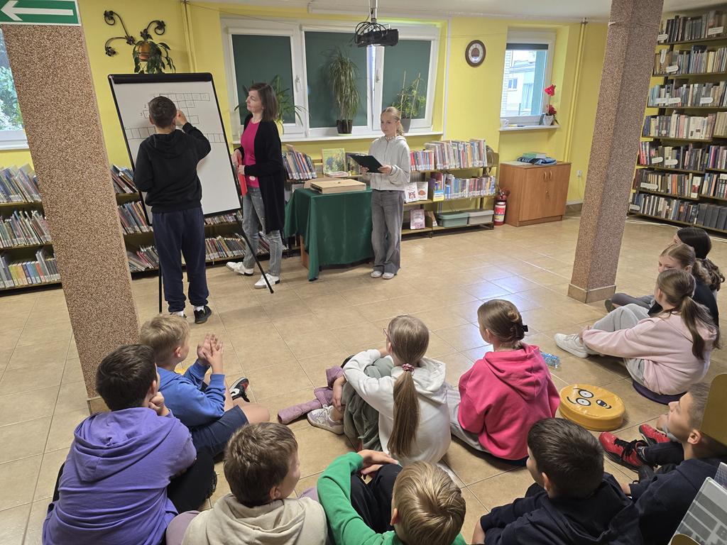 Nauczyciel bibliotekarz w czarnym swetrze i różowej bluzce, niebieskich jeansach stoi przy tablicy do pisania. Przy tablicy stoi też chłopiec na czarno ubrane i pisze. Obok Bibliotekarza stoi dziewczyna w jasnej bluzie i spodniach. Dziewczyna czyta zagadki, za nią stoi stolik z zielonym obrusem. Na przeciwko tych trzech osób siedzi grupa dzieci. W tle widać regały z książkami.