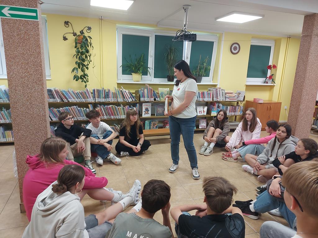 Grupa dzieci 12/13-letnich siedzi w kręgu na podłodze i patrzy na prowadzącą zajęcia. Prowadząca stoi w środku kręgu i pokazuje dzieciom otwartą książkę.