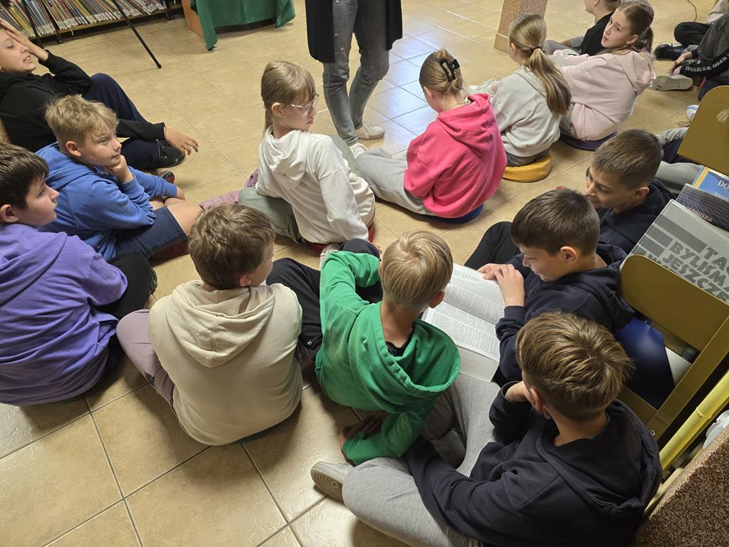Grupa dzieci w kolorowych ubraniach siedzi na siedziskach. Jeden z uczniów szuka w słowniku języka polskiego słowa "współpraca".