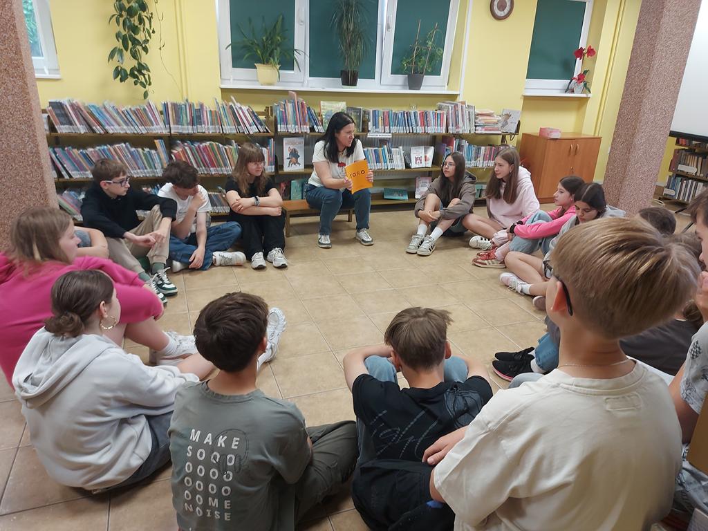Grupa dzieci 12/13-letnich siedzi w kręgu na podłodze i patrzy na prowadzącą zajęcia. Ta również siedzi w kręgu. Jest ubrana w białą podkoszulkę z motywem Muminków i trzyma w dłoni pomarańczową książkę zatytułowaną „TORO”.