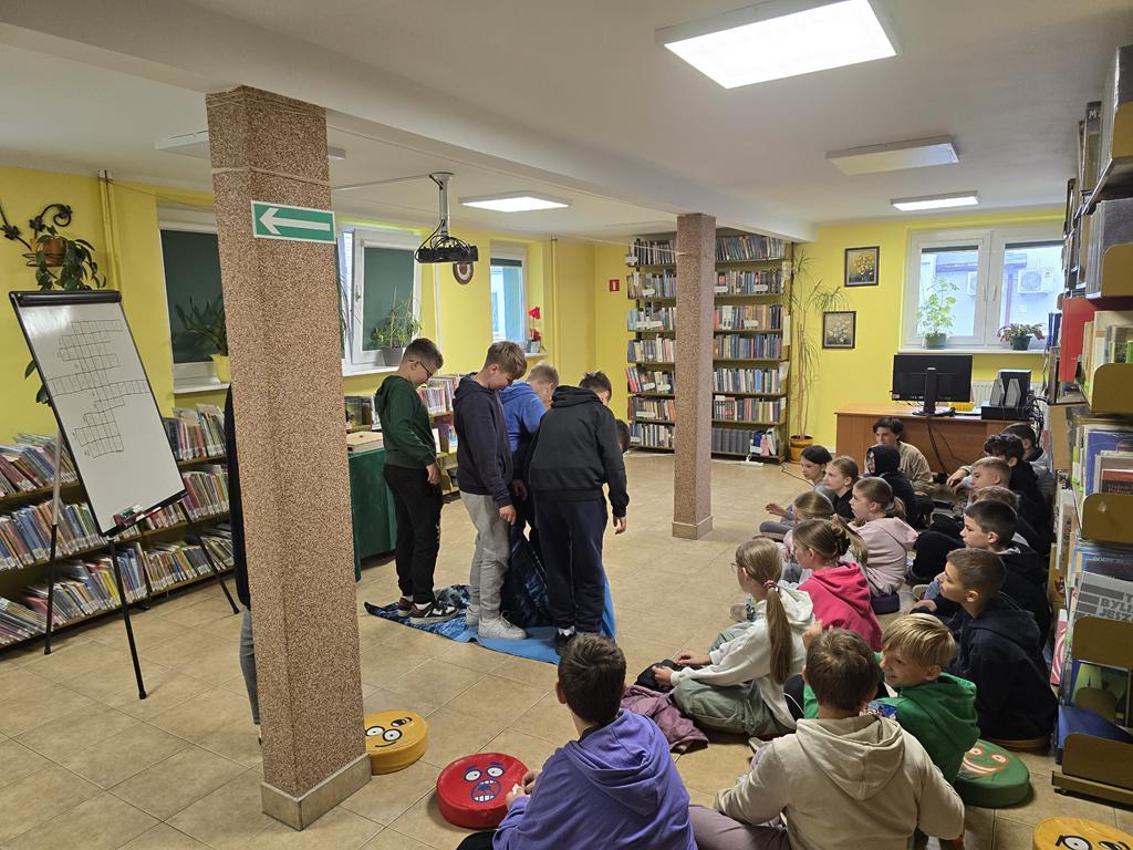 Grupa dzieci w kolorowych ubrania siedzi na siedziskach. Przed nimi leży niebiesko-granatowy kocyk, na którym stoi pięciu chłopców. Z prawej strony od chłopców jest tablica do pisania z krzyżówką. W tle widać regały z książkami.