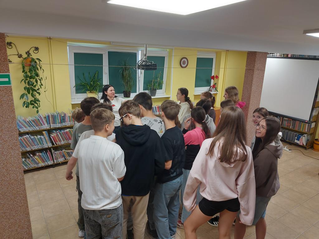 Grupa dzieci 12/13-letnich zbiła się przed prowadzącą zajęcia w ścisłą gromadę. W tle regały z książkami.