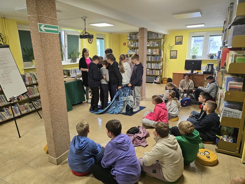 Grupa dzieci w kolorowych ubrania siedzi na siedziskach. Przed nimi leży niebiesko-granatowy kocyk, na którym stoi ośmioro dzieci, próbujących przełożyć kocyk na lewą stroję, bez schodzenia z kocyka. Za dziećmi stoi bibliotekarz. Po lewej stronie stoi tablica do pisani. W tle widać regały z książkami.