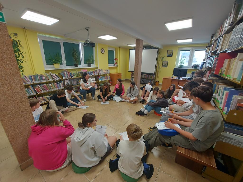 Grupa dzieci 12/13-letnich siedzi w kręgu na podłodze i patrzy na prowadzącą zajęcia. Ta również siedzi w kręgu. Jest ubrana w białą podkoszulkę z motywem Muminków. Siedzą w oświetlonym pomieszczeniu z oknami częściowo zasłoniętymi zielonymi roletami. W pomieszczeniu znajduje się dużo regałów z książkami. Dzieci trzymają w dłoniach kartki.