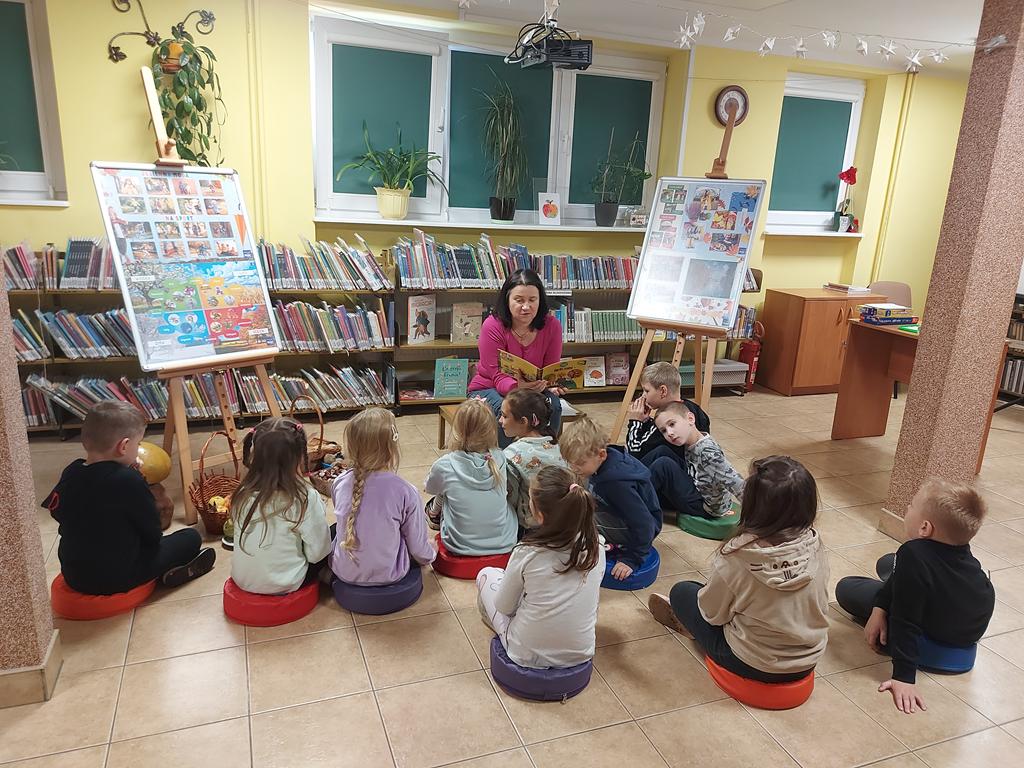 Kolorowo wyposażona sala. Na niskim stołeczku na środku siedzi kobieta w różowej bluzce. Twarz ma zwróconą do przodu. Po jej prawej stronie stoi sztaluga, a na niej jesienne ilustracje. Po jej lewej stronie stoi podobna sztaluga, a pod nią w koszyczkach i pudełkach leżą warzywa i owoce. Między dwiema kolumnami, tyłem, siedzi na podłodze jedenaścioro sześcioletnich dzieci. Patrzą na kobietę.