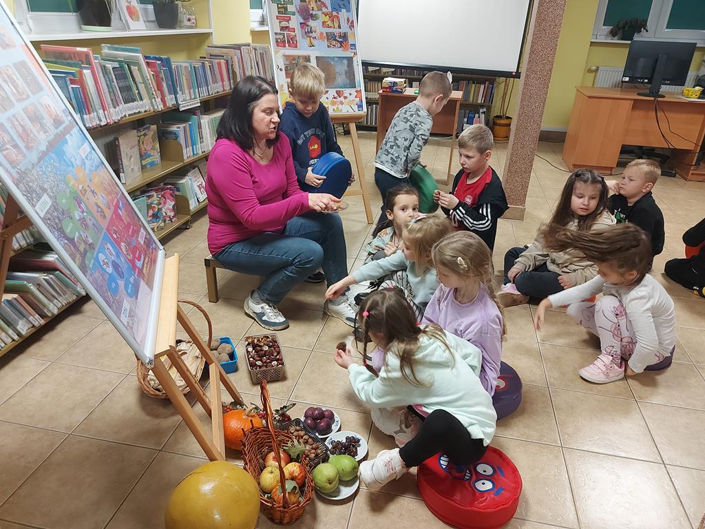 Kolorowo wyposażona sala. Na niskim stołeczku po lewej stronie siedzi bokiem kobieta w różowej bluzce. W ręce trzyma talerzyk z orzechami włoskimi. Jedenaścioro dzieci, siedzących na podłodze ogląda i dotyka jesienne owoce i warzywa.