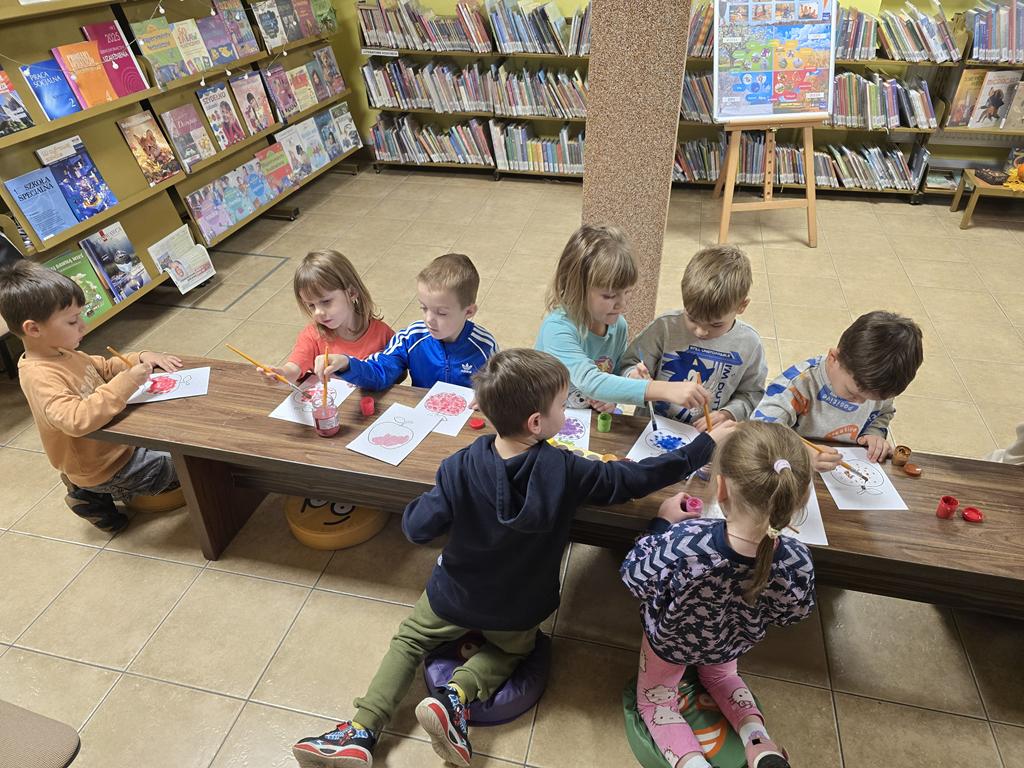 Ośmioro dzieci w kolorowych ubrania siedzą przy ławce. Malują farbami jabłka. W tle są regały z książkami.