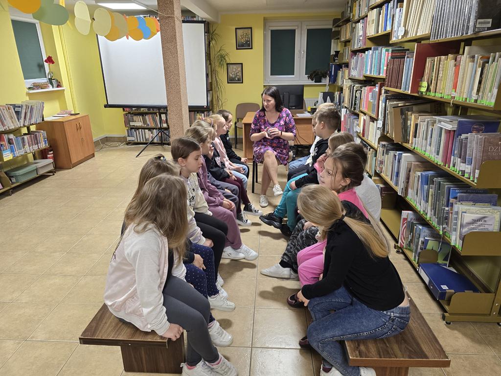 Piętnaścioro dzieci siedzi na dwóch podłużnych niskich ławkach. Twarzami dzieci są zwrócone do siebie. Kobieta siedzi z tyłu pomiędzy dwoma ławkami, wśród dzieci, i trzyma w dłoniach kłębek kolorowej włóczki.