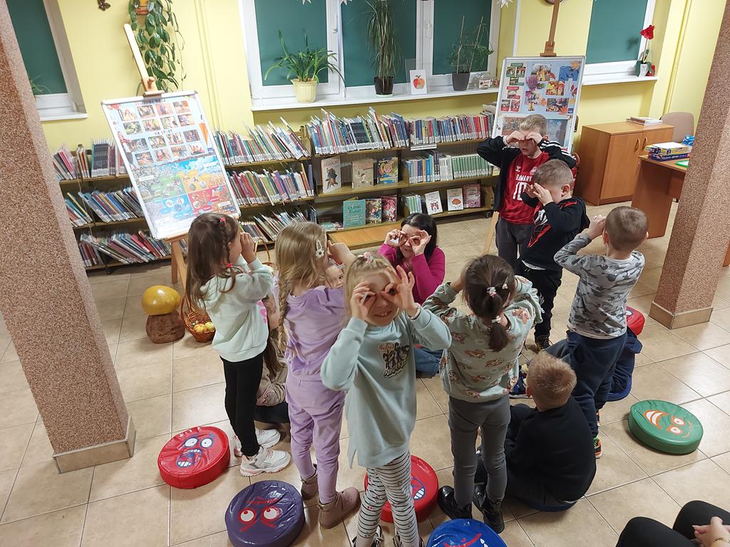 Dzieci siedzą w kółeczku na podłodze. Cztery dziewczynki i trzech chłopców stoją i patrzą przez lornetki zrobione z dłoni. Kobieta, siedząca na podłodze też patrzy przez lornetkę z dłoni.