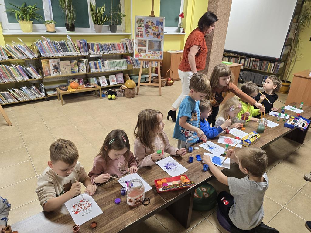 Ośmioro dzieci w kolorowych ubrania siedzą przy ławce. Malują farbami jabłka. Obok nich pochylona sto kobieta w pomarańczowej bluzce i białych spodniach. W tle są regały z książkami, ekran, tablica z plakatami dotyczącymi jesieni, grzyb, kosze, ławeczka.