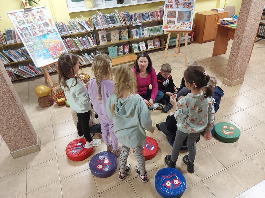 Dzieci siedzą w kółeczku na podłodze. Cztery dziewczynki stoją i patrzą na kobietę, która siedzi na podłodze i patrzy na dziewczynki. Kobieta ma otwarte usta.