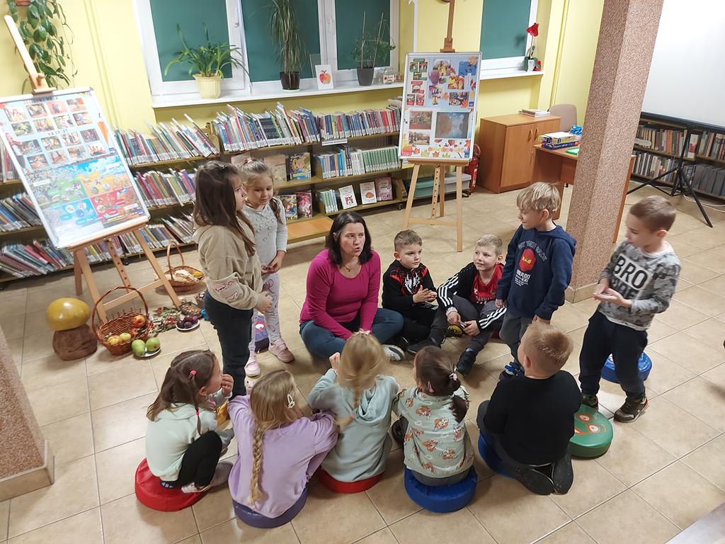 Kolorowo wyposażona sala. Na poduszce na środku siedzi kobieta w różowej bluzce. Twarz ma zwróconą do przodu, ma otwarte usta. Po jej prawej stronie stoi sztaluga, a na niej jesienne ilustracje. Po jej lewej stronie stoi podobna sztaluga, a pod nią w koszyczkach i pudełkach leżą warzywa i owoce. Między dwiema kolumnami, tyłem, siedzą na podłodze. Po lewej stronie stoi dwóch chłopców. Po prawej stronie stoją dwie dziewczynki.