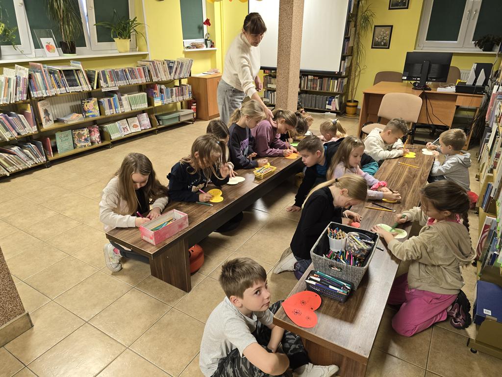 Piętnaścioro dzieci siedzi na podłodze na kolorowych siedziskach. Pochylają się nad dwoma niskimi długimi stolikami. Dzieci mają przed sobą kolorowe papierowe serca, w których piszą ołówkami. Po lewej stronie kobieta w białej bluzce i dżinsowych spodniach schyla się nad ławką i wyciąga prawą dłoń w stronę dzieci.