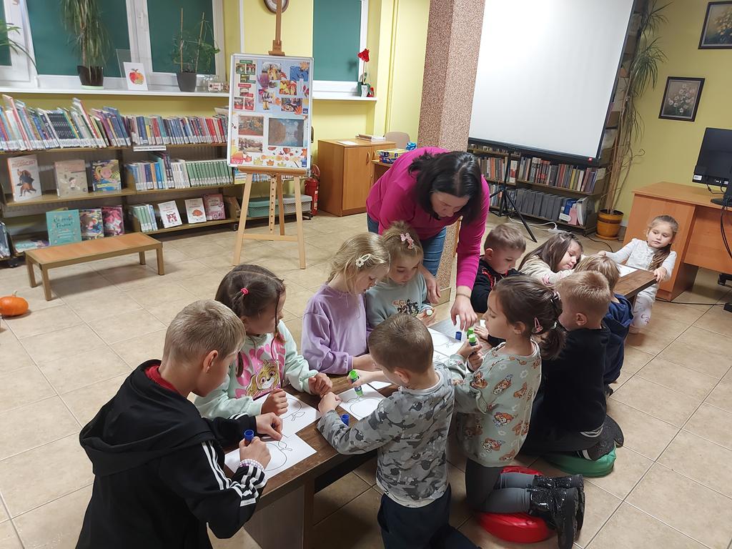 Jedenaścioro dzieci siedzi na podłodze wokół niskiego podłużnego stolika. Nad dziećmi pochyla się kobieta. Na stoliku leżą kartki z rysunkami jabłek. Dzieci smarują klejem narysowane owoce.