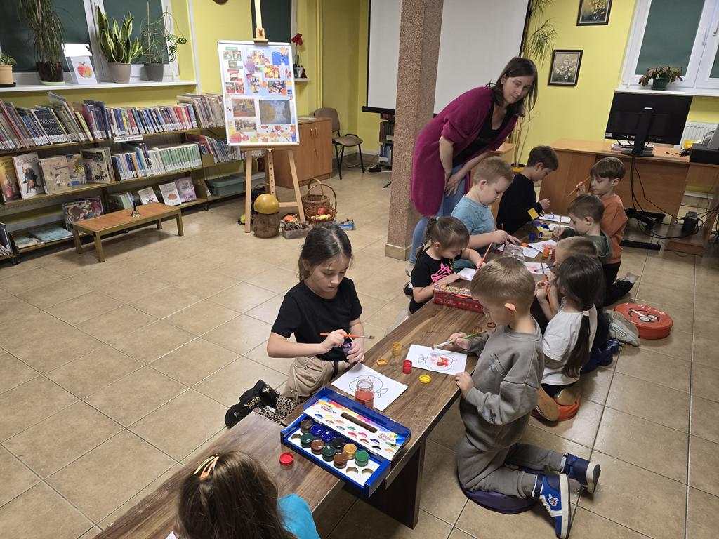 Dziesięcioro dzieci w kolorowych ubrania siedzą przy ławce. Malują farbami jabłka. Obok nich pochylona sto kobieta w ciemnym różowym swetrze, niebieskich spodniach. W tle są regały z książkami, ekran, tablica z plakatami dotyczącymi jesieni, grzyb, kosze, ławeczka.