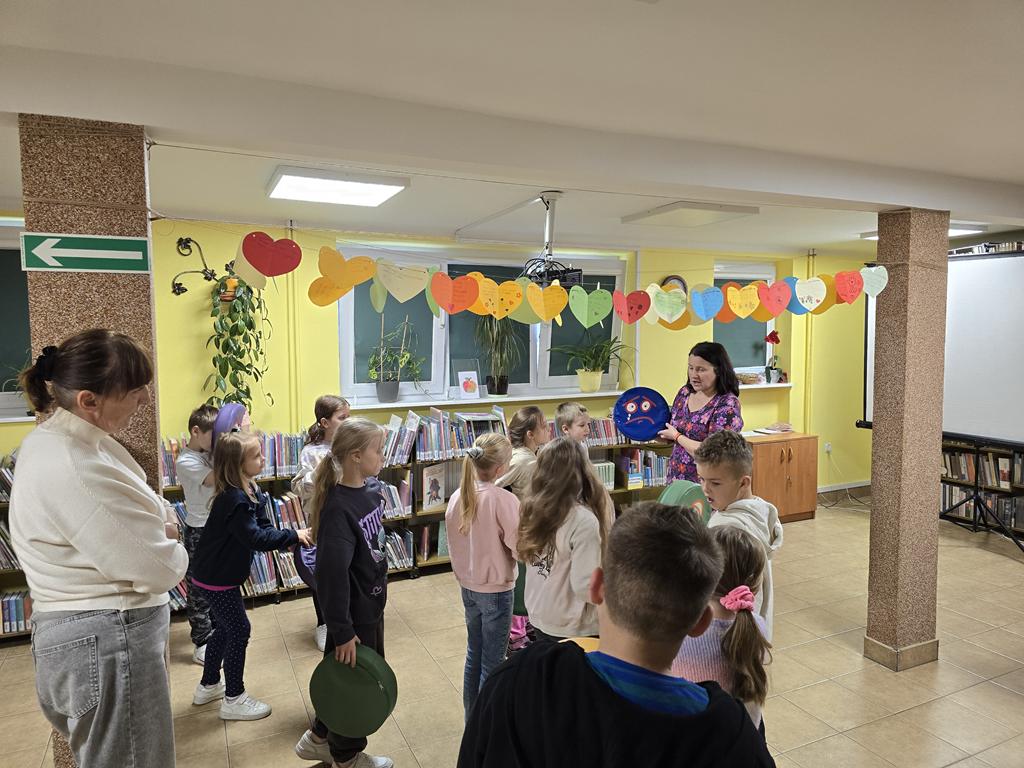 Kobieta stoi zwrócona w lewą stronę. Trzyma w rękach okrągłą poduszkę z naklejoną smutną miną. Wokół niej stoi w rozproszeniu grupa jedenaściorga dzieci zwróconych w stronę kobiety. Nad głowami dzieci między dwoma kolumnami jest rozwieszona nić z kolorowymi papierowymi sercami. Po lewej stronie przy kolumnie stoi kobieta w jasnej bluzce i dżinsowych spodniach. Patrzy na dzieci.