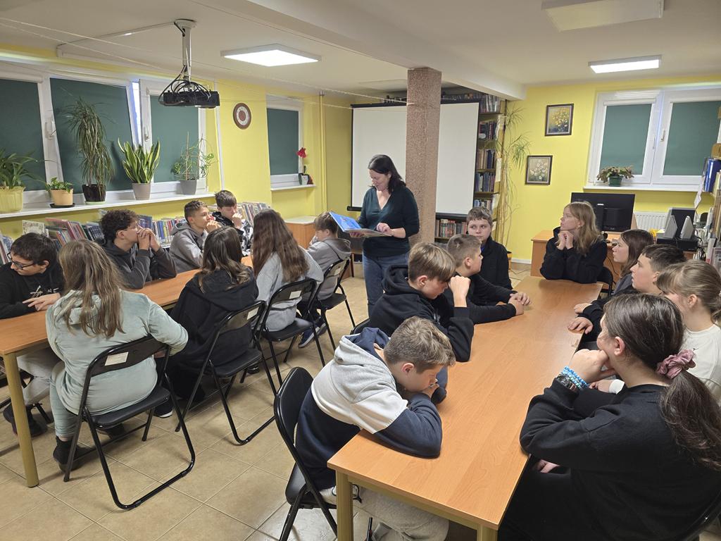 Kobieta ubrana na zielono stoi z pochyloną głową między dwiema grupami uczniów, trzyma w ręku otwartą książkę. Trzynastoletnie dzieci siedzą przy podłużnych stolikach na rozkładanych krzesłach. Po lewej stronie siedzi pięciu chłopców i trzy dziewczynki, po prawej siedzi pięciu chłopców i cztery dziewczynki.