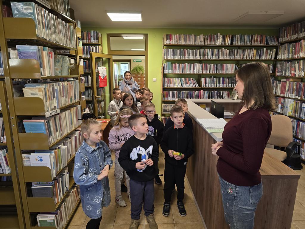 Grupa dzieci w kolorowych ubrania, oraz pani nauczycielka wchodzą do wypożyczalni. Przed nimi obok lady bibliotecznej stoi kobieta z ciemno-bordowym swetrze i niebieskich spodniach. Po prawej i lewej stronie są regały z książkami.