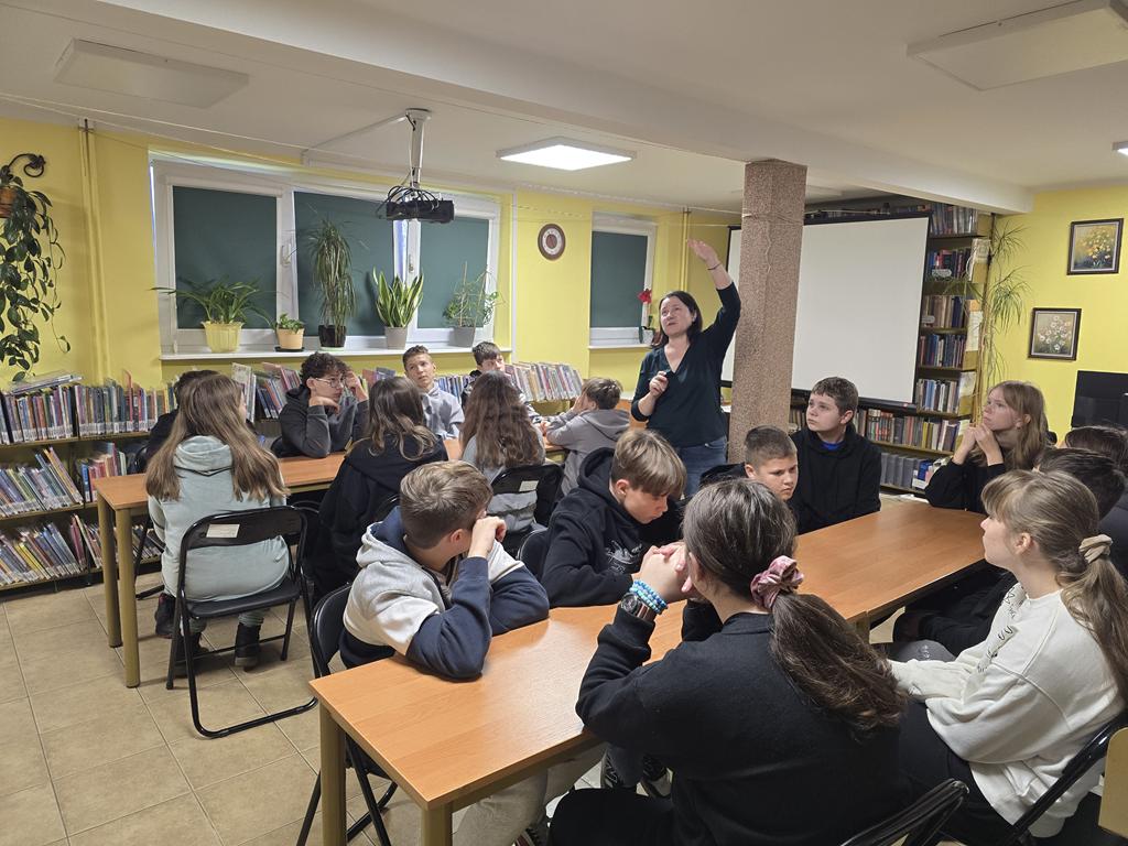 Kobieta ubrana na zielono stoi z podniesioną do góry ręką między dwiema grupami uczniów. Trzynastoletnie dzieci siedzą przy podłużnych stolikach na rozkładanych krzesłach. Po lewej stronie siedzi pięciu chłopców i trzy dziewczynki, po prawej siedzi pięciu chłopców i cztery dziewczynki.