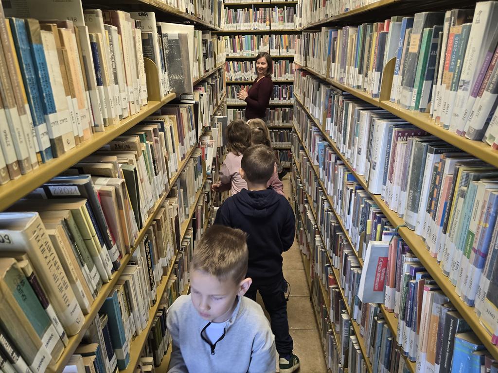 Pięcioro dzieci chodzi między regałami z książkami. Na końcu alejki stoi kobieta w ciemno -bordowym swetrze.