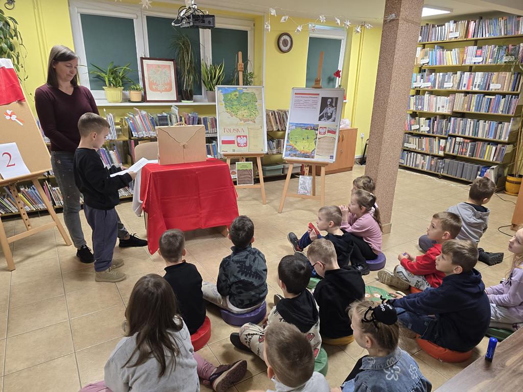 Kobieta w ciemno-bordowym swetrze i niebieskim spodniach stoi obok chłopca w ciemnych ubraniach, czytającego z białej kartki zagadkę. Obok nich stoi stolik z obrusem biało-czerwonym, na którym stoi teatrzyk kamishibai. Obok stolika stoją dwie tablice z plakatami dotyczącymi Narodowego Święta Niepodległości.
Przed kobietą i chłopcem siedzi grupa dzieci w kolorowych ubraniach. Na parapecie stoi godło Polski. W tle widać regały z książkami.