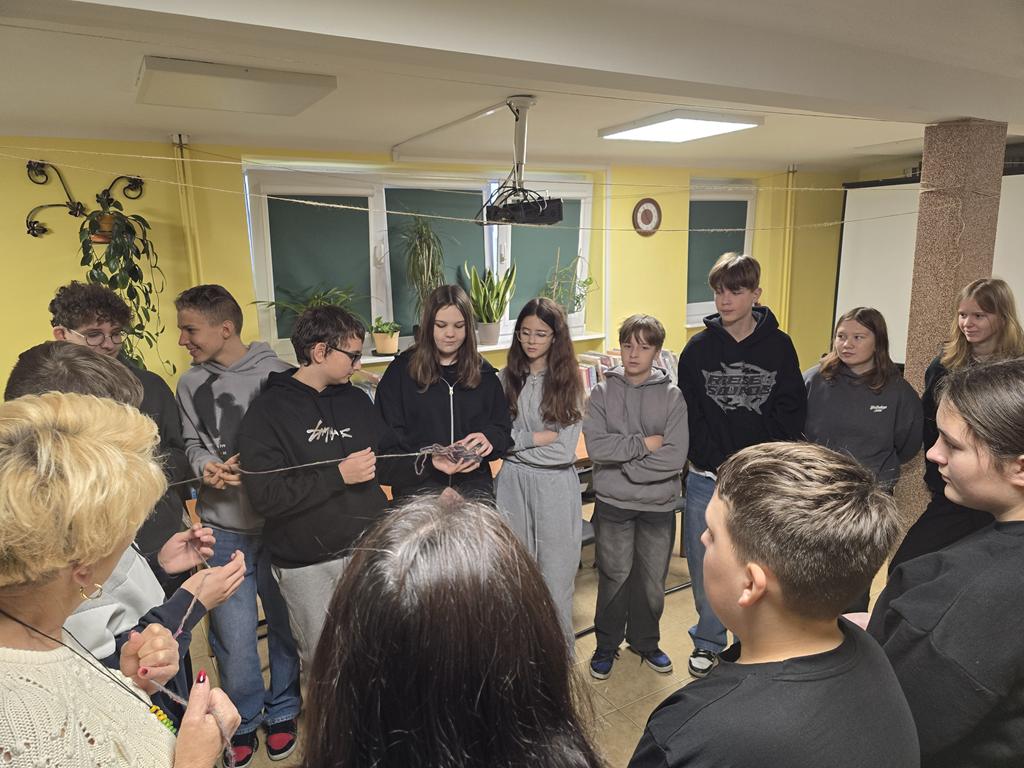 Dwie kobiety i dzieci stoją w kręgu na środku sali. Trzymają w ręku nić z rozwiniętego kłębka włóczki.