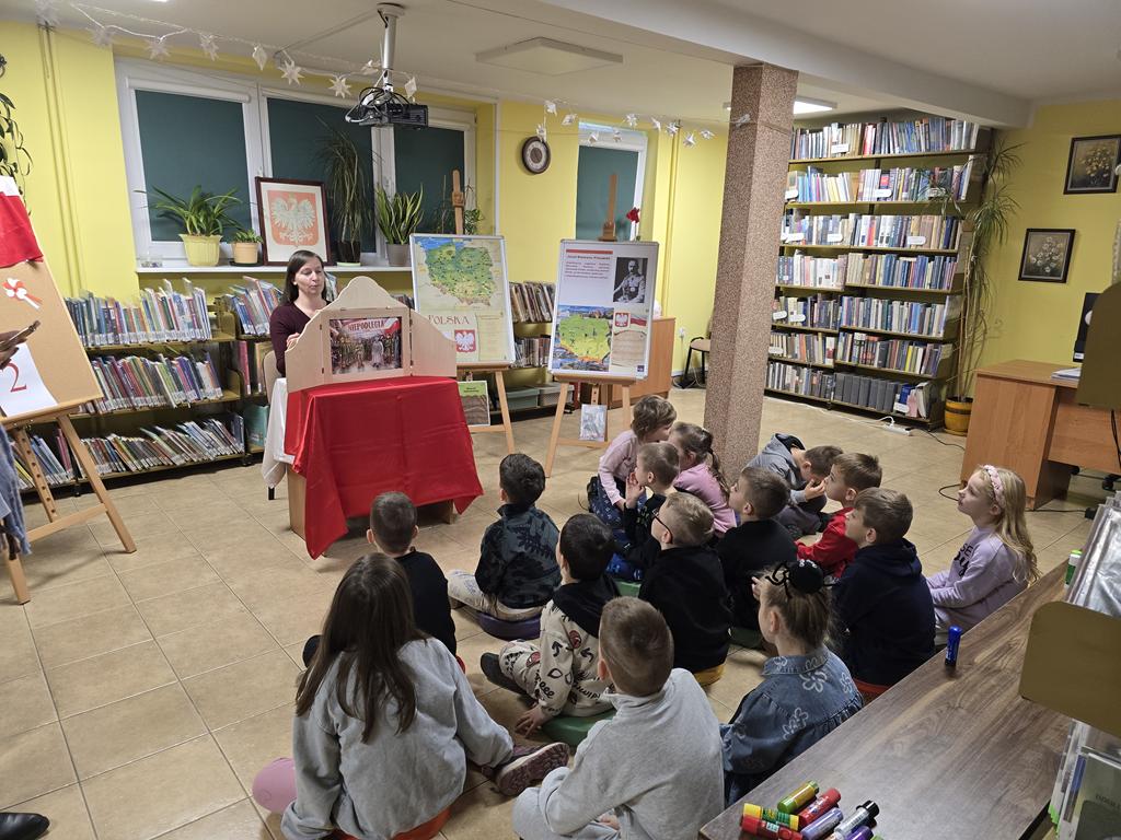 Kobieta w ciemno-bordowym swetrze siedzi za stolikiem z obrusem biało-czerwonym, czyta opowiadanie z teatrzyku kamishibai pod tytułem "Niepodległa 11 listopada". Po prawej stronie kobiety stoją dwie tablice z plakatami dotyczącymi Narodowego Święta Niepodległości. Przed kobietą siedzi grupa dzieci w kolorowych ubraniach. Za nimi jest brązowa ławka, na której leżą kleje. Na parapecie stoi godło Polski. W tle widać regały z książkami.