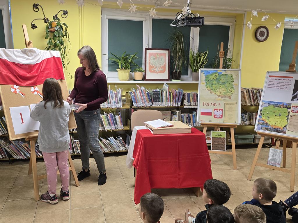 Kobieta w ciemno-bordowym swetrze i niebieskim spodniach stoi obok dziewczynki w szarej bluzie i różowych spodniach. Dziewczynka przyczepia kartki do tablicy. Na tablicy jest flaga Polski, kotyliony. Obok nich stoi stolik z obrusem biało-czerwonym, na którym leży teatrzyk kamishibai. Obok stolika stoją dwie tablice z plakatami dotyczącymi Narodowego Święta Niepodległości. Na parapecie stoi godło Polski. Przed kobieta i dziewczynką siedzi grupa dzieci w kolorowych ubraniach. W tle widać regały z książkami.