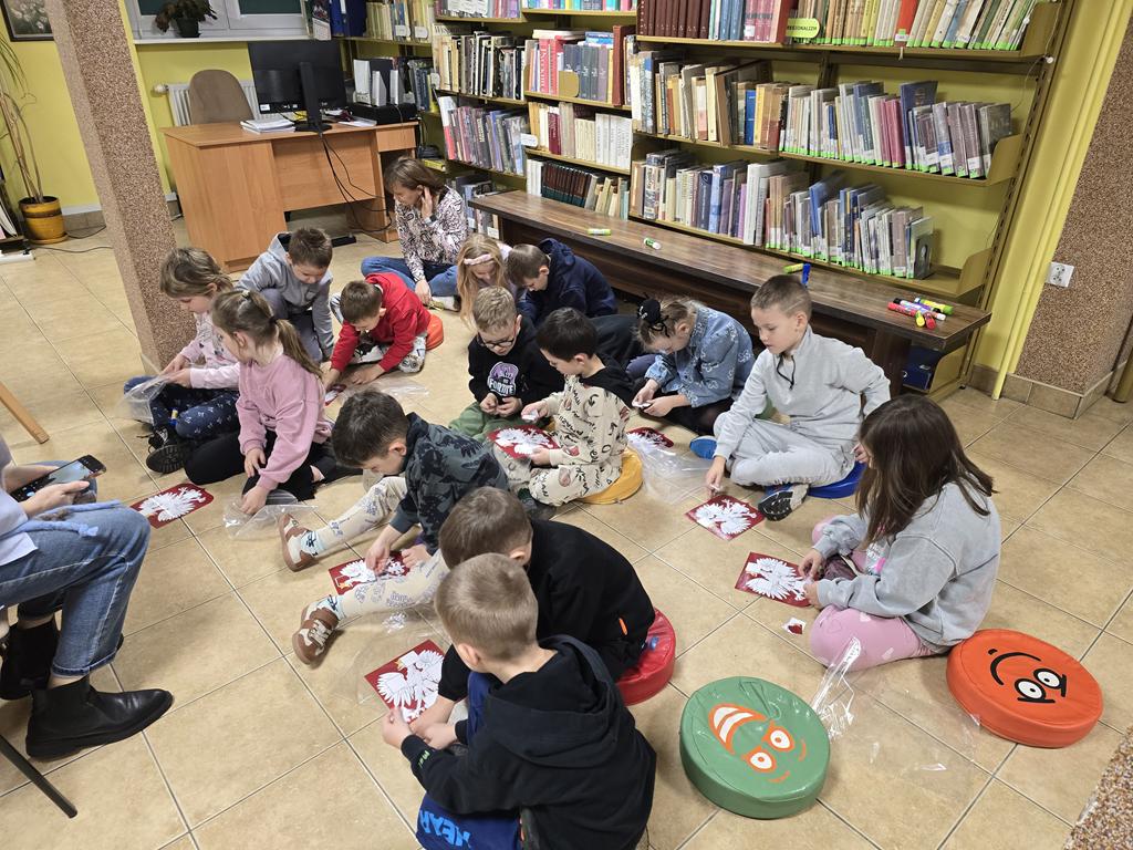 Grupa dzieci ubrana na kolorowo siedzi na emotikonowych, kolorowych pufach, układa z puzzli godło. Za dziećmi są regały z książkami. Po lewej stroni stoi burko.