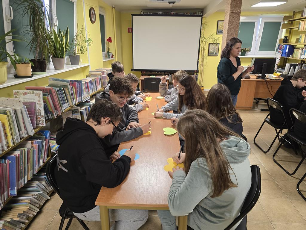 Ośmioro dzieci siedzi przy stoliku i pisze na kolorowych papierowych sercach. Po prawej stronie stoi kobieta ubrana na zielono.