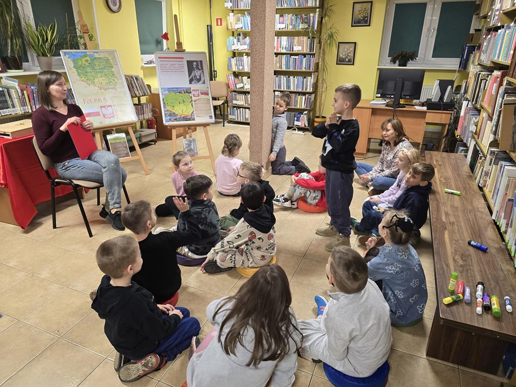 Kobieta w ciemno-bordowym swetrze i niebieskim spodniach siedzi na krześle na środku sali, trzyma czerwoną podkładkę. Po lewej stronie kobiety są dwie tablice z plakatami dotyczącymi Narodowego Święta Niepodległości. Za kobietą jest stolik z obrusem biało-czerwonym i regały z książkami. Przed kobietą siedzi grupa dzieci w kolorowych ubraniach. Jeden chłopiec w ciemnych ubraniach stoi. Za dziećmi stoi brązowa ławka (na niej kleje), a za nią regały z książkami.