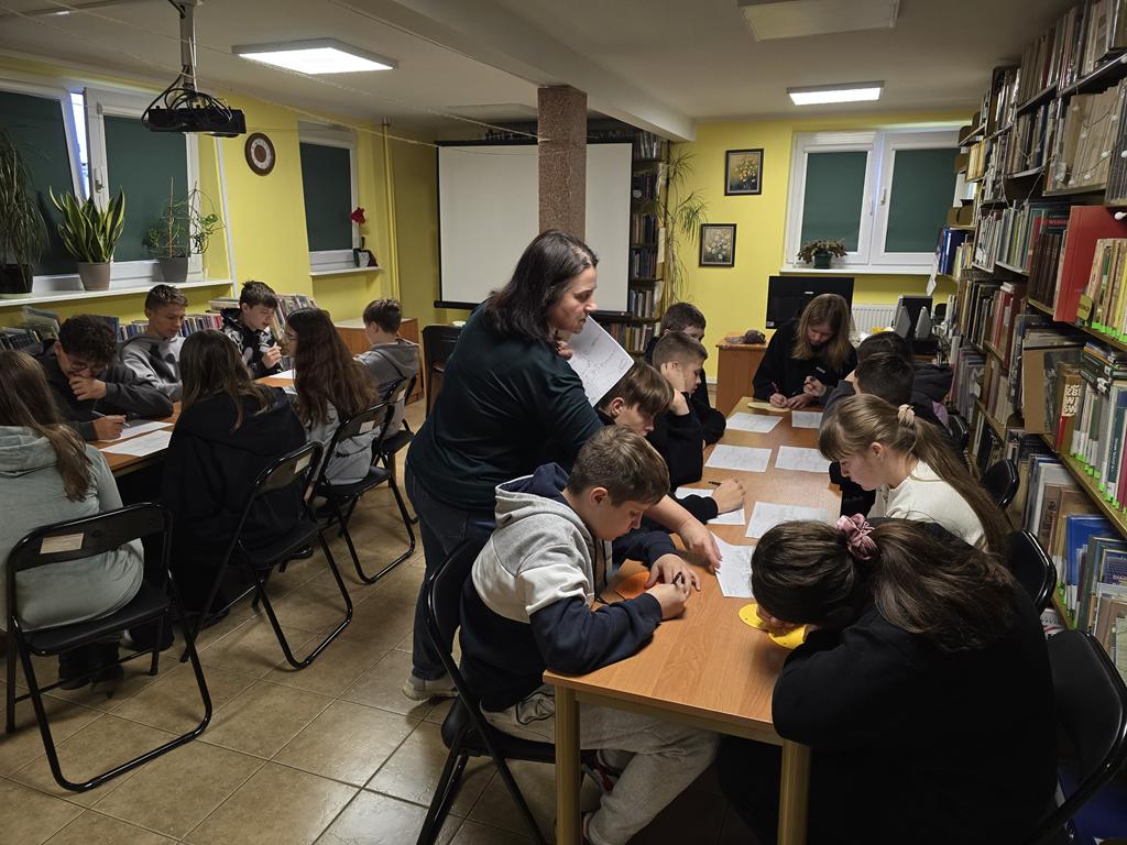 Przy dwóch stolikach siedzą dzieci. Kobieta stojąca pomiędzy nimi rozdaje kartki z zadaniami.