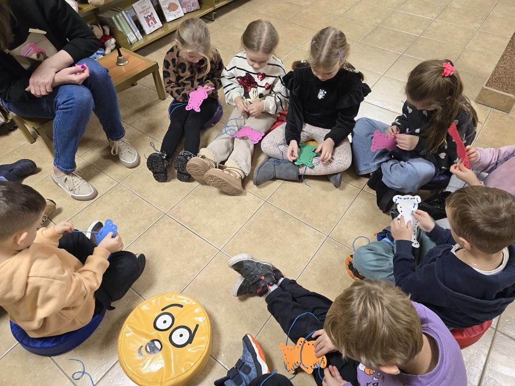 Dzieci w kolorowych ubraniach siedzą w kole. Każde ma misia z pianki dekoracyjnej. Dzieci robią misiowe wyszywanki bez igły.  