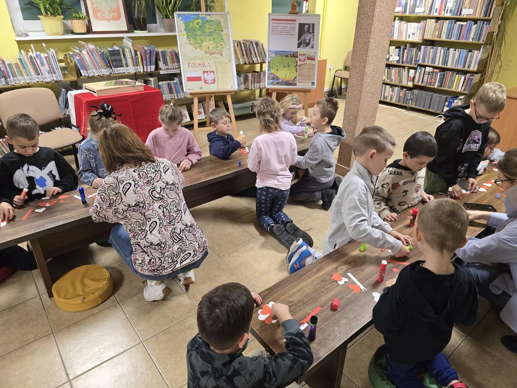 Grupa dzieci w kolorowych ubraniach i dwie kobiety siedzą przy dwóch ławkach i wykonują kotyliony. Na ławkach są elementy kotyliony i kleje. Za dziećmi jest stolik z obrusem biało-czerwonym, dwie tablice z plakatami związanymi z Narodowym Świętem Niepodległości. Na parapecie stoi godło Polski. W tle są regały z książkami.