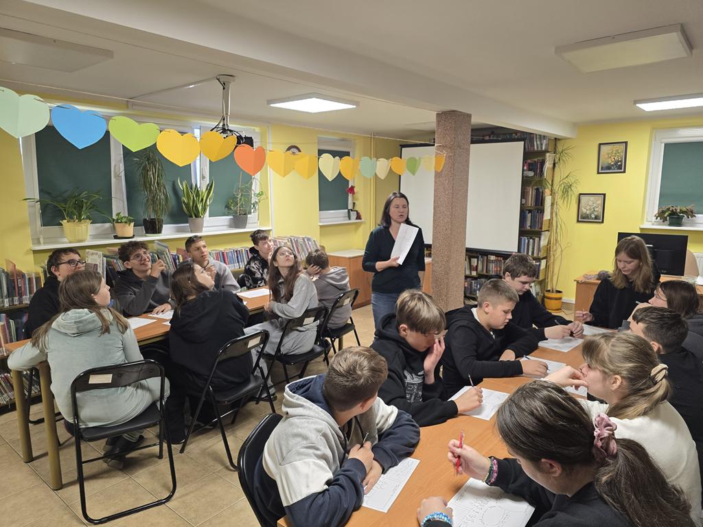 Przy dwóch stolikach siedzą dzieci. Kobieta stojąca pomiędzy nimi trzyma w ręku kartkę z zadaniami. Nad nią wisi girlanda z kolorowych papierowych serc. Na stolikach przed dziećmi leżą wypełnione kartki z zadaniami.