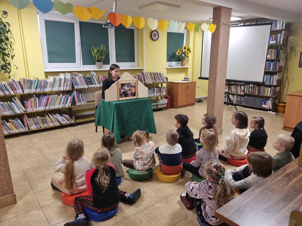 Bibliotekarka w czarnej bluzce siedzi przy stoliku z zielonym obrusem. Na stoliku teatrzyk kamishibai, który czyta kobieta. Grupa dzieci w kolorowych ubrania siedzi przed nią i słucha opowiadania. Za bibliotekarką są regały z książkami. Po jej prawej stronie jest biały ekran do rzutnika.