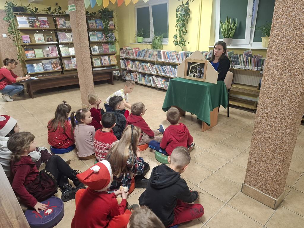 Bibliotekarka w czarnym swetrze i niebieskiej bluzce siedzi przy stoliku z zielonym obrusem. Na stoliku teatrzyk kamishibai, który czyta kobieta. Grupa dzieci w kolorowych ubrania siedzi przed nią i słucha opowiadania. Za bibliotekarką są regały z książkami.