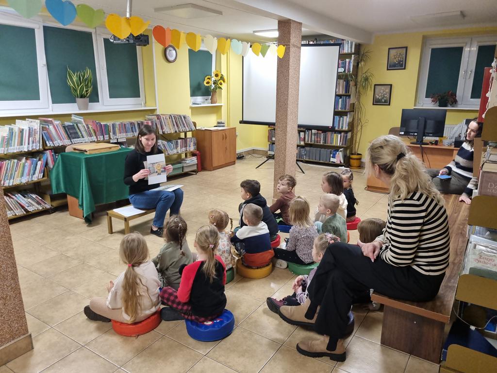 Bibliotekarka w czarnej bluzce i niebieskich spodniach siedzi na ławeczce i pokazuje dzieciom rysunki. Za kobietą jest stolik z zielonym obrusem i regały z książkami. Grupa dzieci w kolorowych ubrania siedzi przed nią i ogląda obrazki. Po prawej stronie kobiety jest biały ekran do rzutnika. Za dziećmi na długiej ławce siedzą dwie panie nauczycielki.