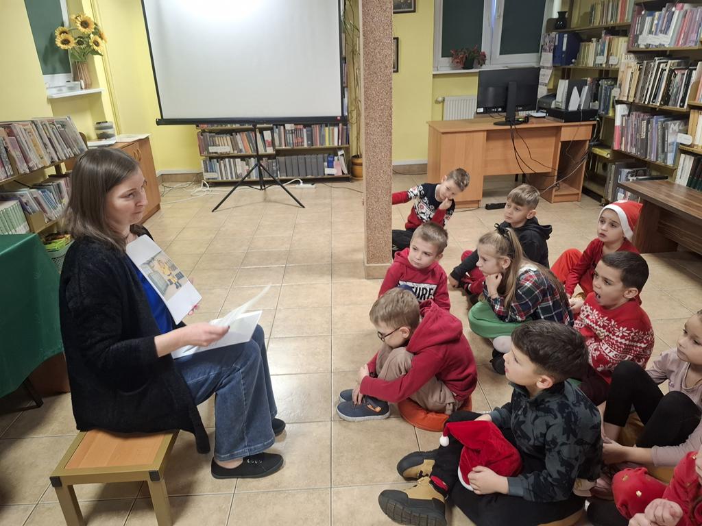 Bibliotekarka w niebieskie bluzce, spodniach, czarnym swetrze siedzi na ławeczce i pokazuje dzieciom rysunki. Za kobietą jest stolik z zielonym obrusem i regały z książkami. Grupa dzieci w kolorowych ubrania siedzi przed nią i ogląda obrazki. W tle jest biały ekran do rzutnika.