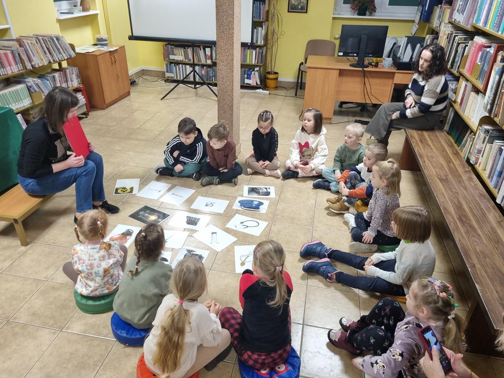 Bibliotekarka w czarnej bluzce i niebieskich spodniach siedzi na ławeczce, ma w ręku czerwoną podkładkę. Grupa dzieci w kolorowych ubrania siedzi w kole. Na środku kola leżąc obrazki związane z pracą górnika. Za dziećmi na długiej ławce siedzi jedna panie nauczycielki.